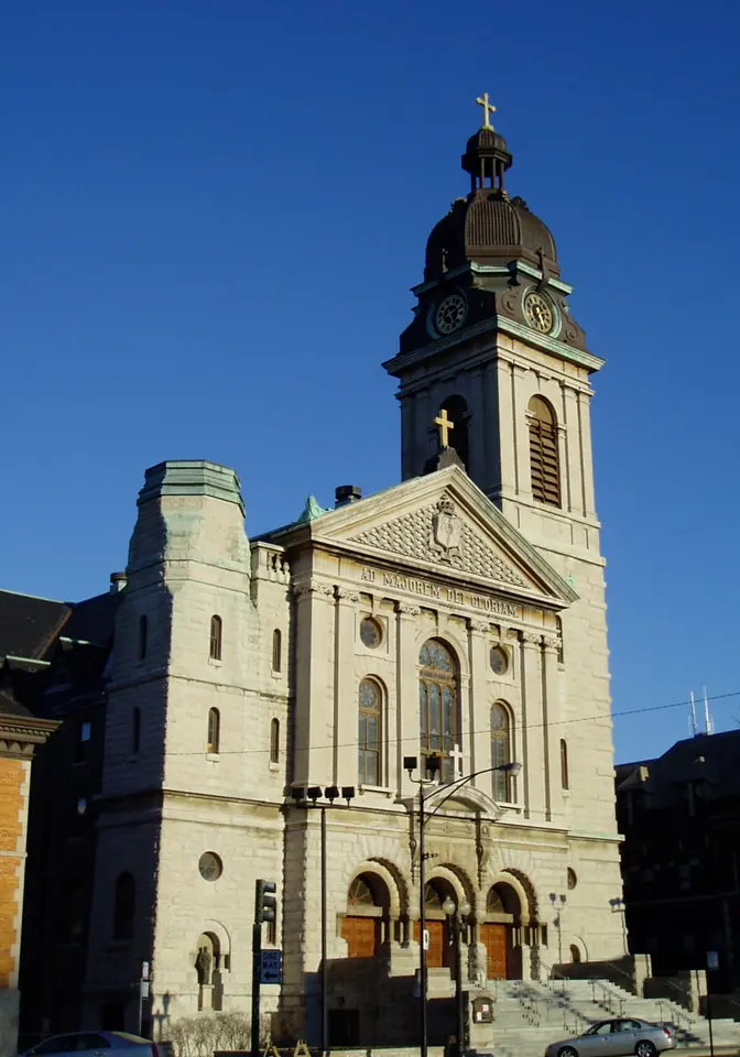 Limestone facade of St. John Cantius Church in Chicago with three arched doors and a tall square clock tower topped by a dome and cross