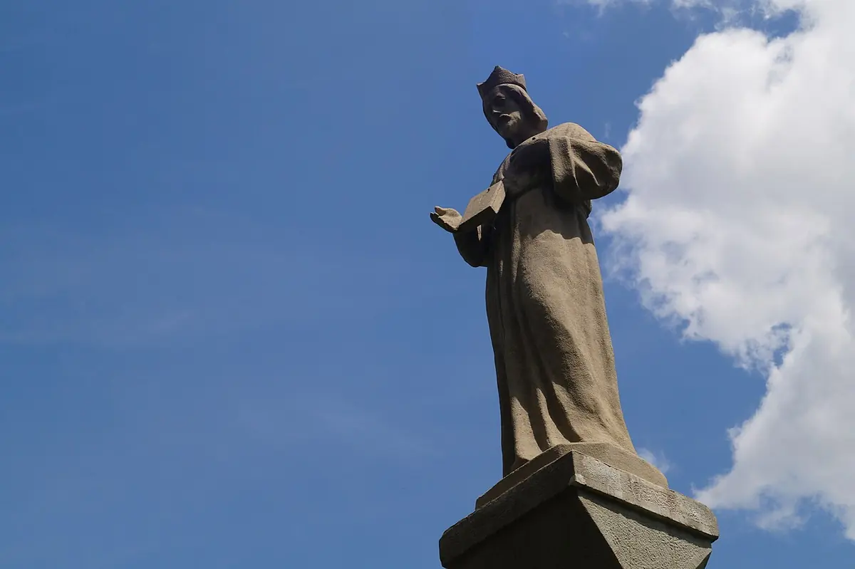 Stone statue of St. John Cantius in a biretta with an open hand, standing on a tall pedestal against a blue sky with white clouds