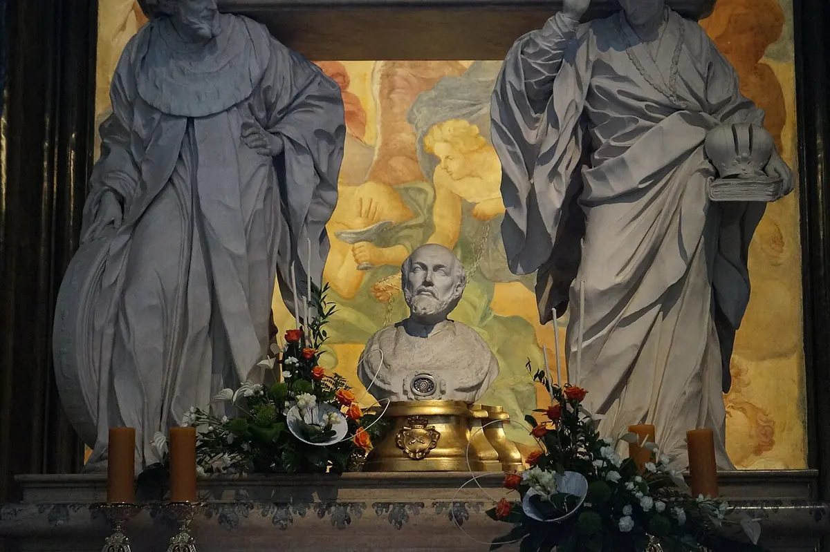 Silver reliquary bust of St. John Cantius on a gilded base between two large gray stone figures, with flowers and candles in front