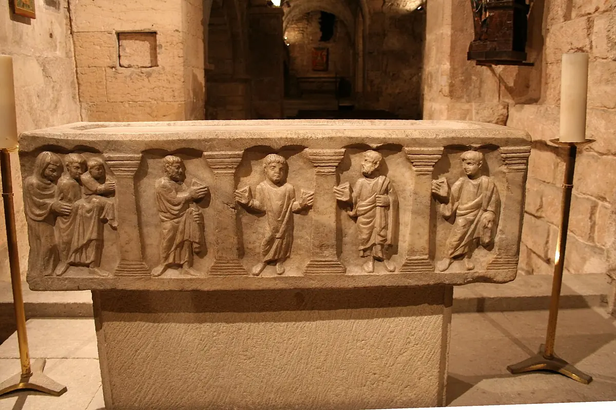 A small carved stone sarcophagus with a row of standing robed figures between columns, in a dim vaulted crypt lit by candles