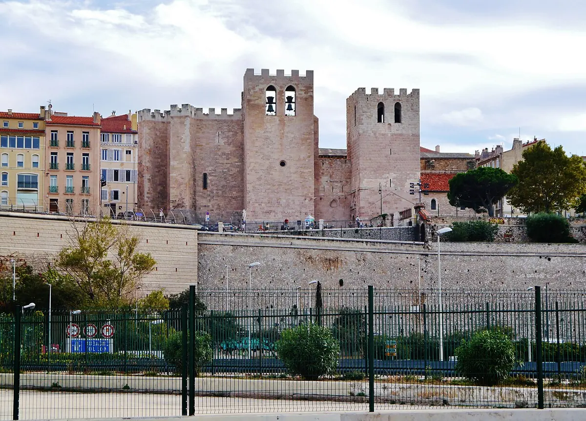 The fortress-like stone abbey church of St. Victor in Marseille with two square crenellated towers rising above a wall and a row of city houses