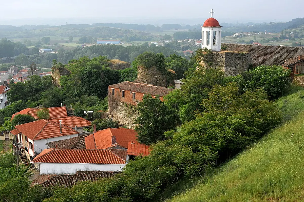 Red-tiled houses and old stone walls climbing a green hillside to a white church bell tower with a red dome