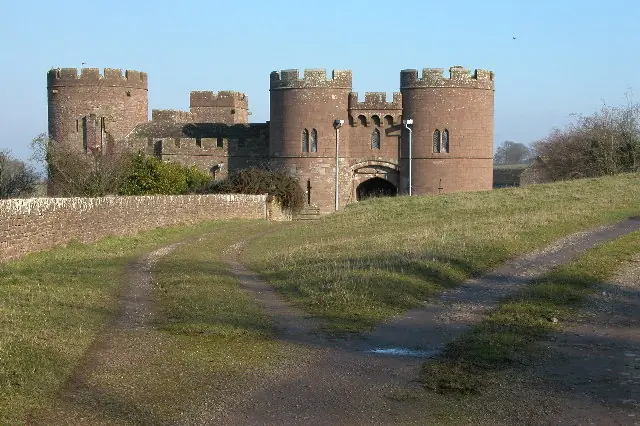 A small red-sandstone castle with round towers and a gatehouse behind a grassy track in the Herefordshire countryside
