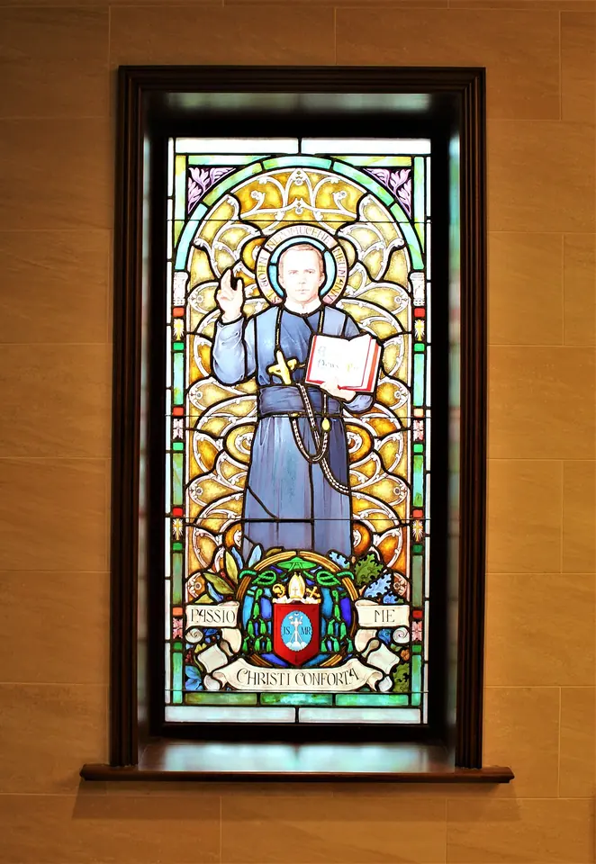 Stained-glass window of St. John Neumann in a dark habit holding a book, above a coat of arms and a banner reading Passio Christi conforta me