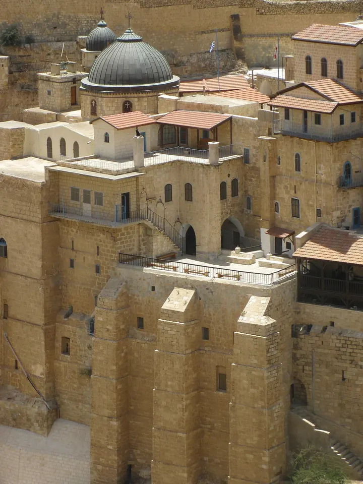 Sand-colored stone buildings of a monastery with blue domes and a bell tower, built in tiers into a cliff face