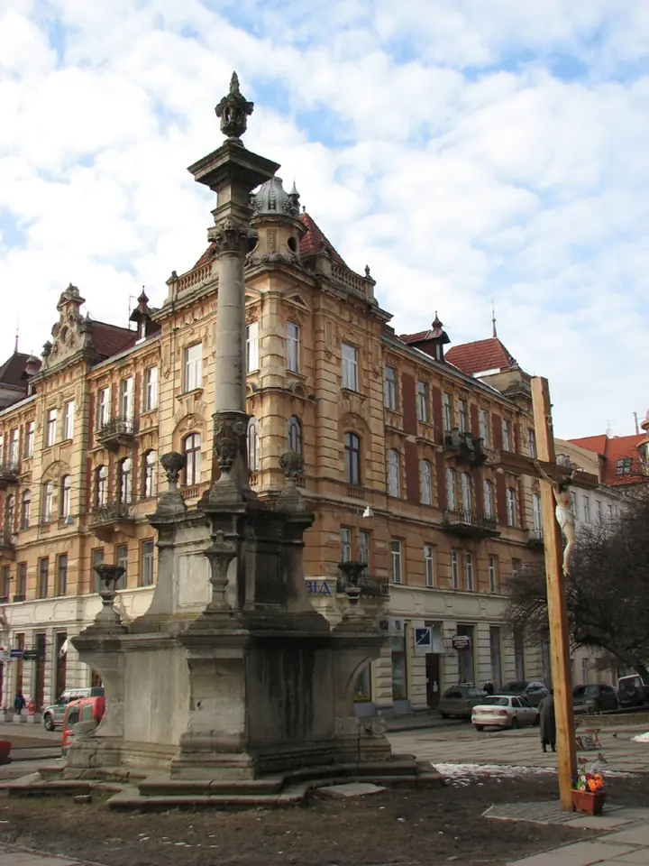 A tall stone column topped with a small statue, standing on a carved base on a city street in front of old apartment buildings