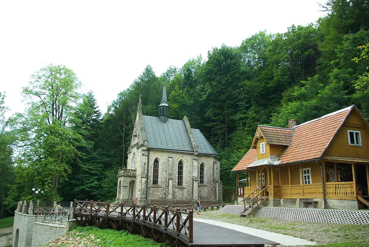 A small gray stone Gothic-revival chapel and a wooden house with a red roof in a forest clearing, reached by a wooden footbridge