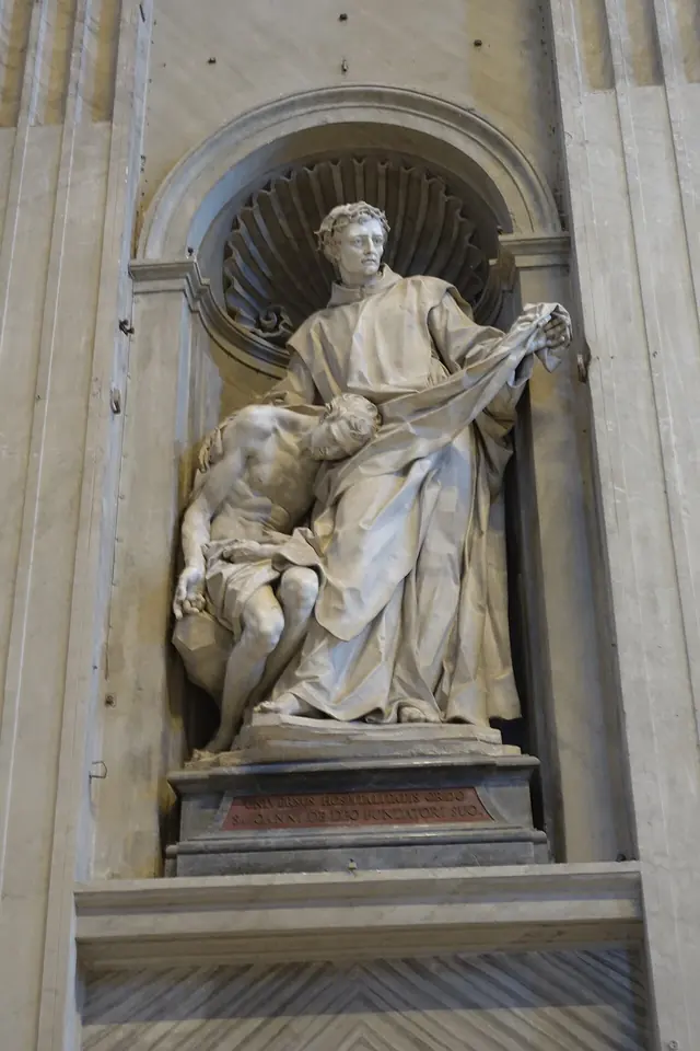A white marble statue in a niche of St. Peter's Basilica showing John of God drawing his cloak around a nearly naked sick man who leans against him