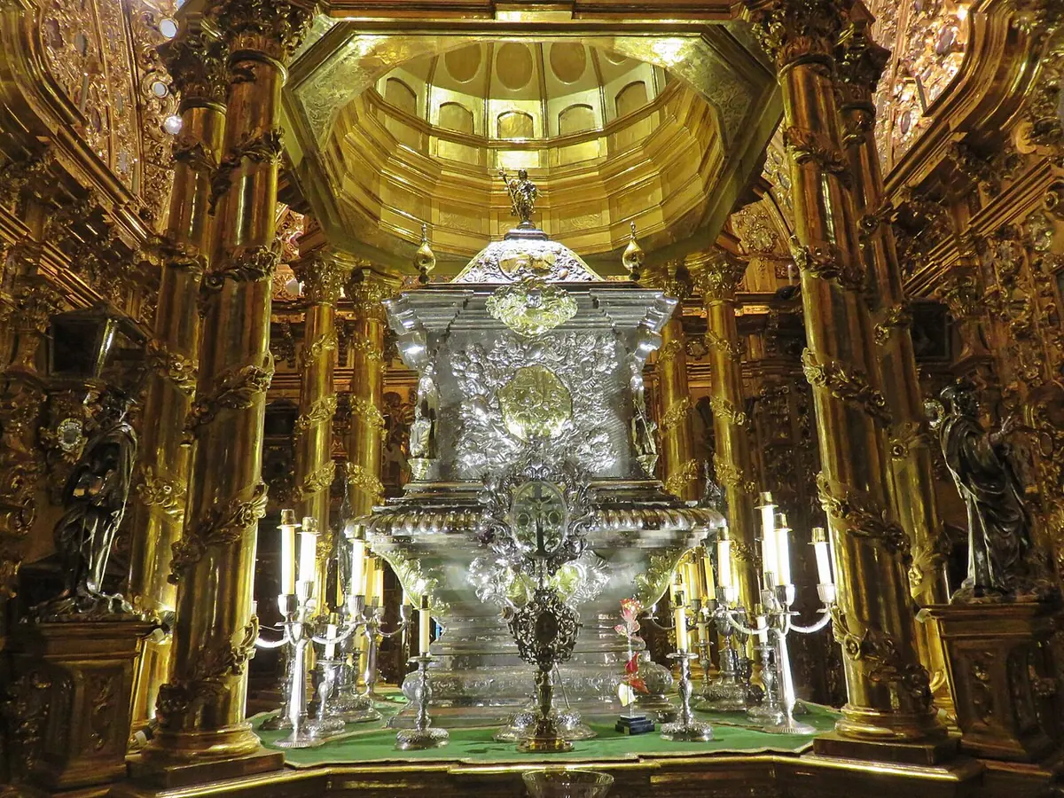 A large embossed silver urn on a pedestal in a small chamber walled with gilded carved columns and mirrors