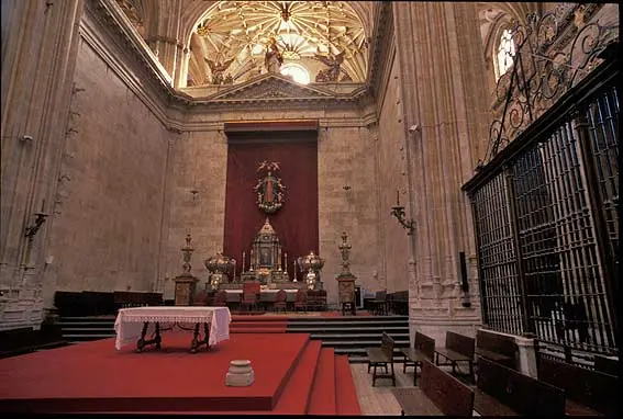 The high altar of a Gothic cathedral with a red hanging behind a statue, silver reliquary urns on the altar steps and an iron grille to the right