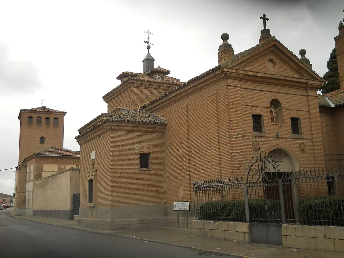 Small brick church with a bell gable and a tiled dome behind an iron fence on a quiet village street