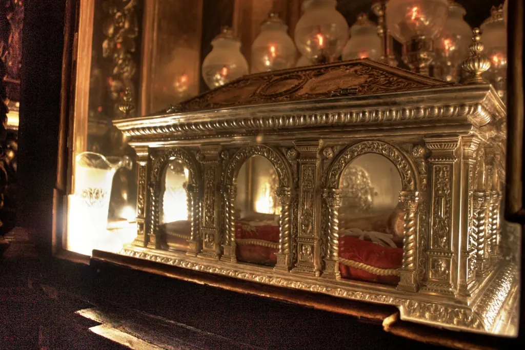 Silver reliquary shaped like a small arcaded chest, with relics wrapped in red visible behind glass, lit by hanging lamps