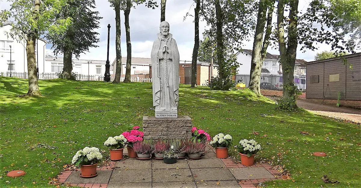 A white statue of a priest holding a crucifix on a pedestal in a grassy park, with pots of pink and white flowers at its base