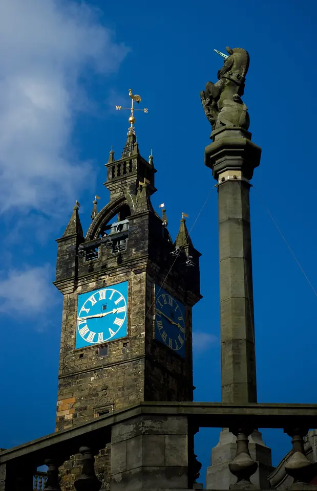 The stone column of a market cross topped by a unicorn beside a dark stone clock tower with a blue clock face, against a deep blue sky
