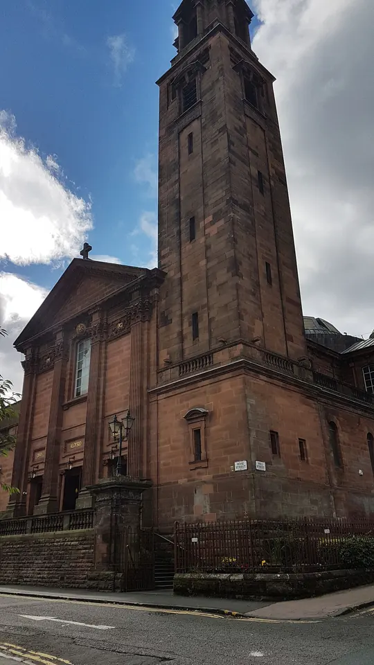 A red sandstone church with a tall square bell tower and arched windows on a city street under a blue sky