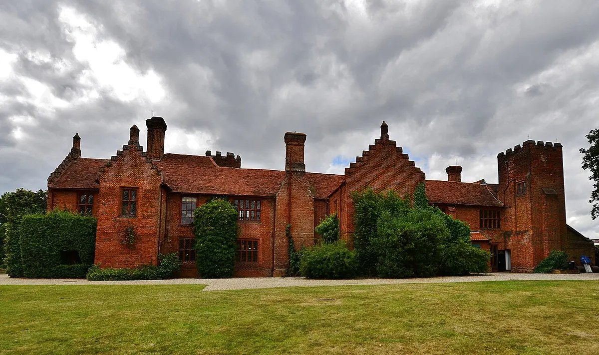 Long red-brick Tudor manor house with stepped gables, tall chimneys and a square tower, behind a wide green lawn under a cloudy sky