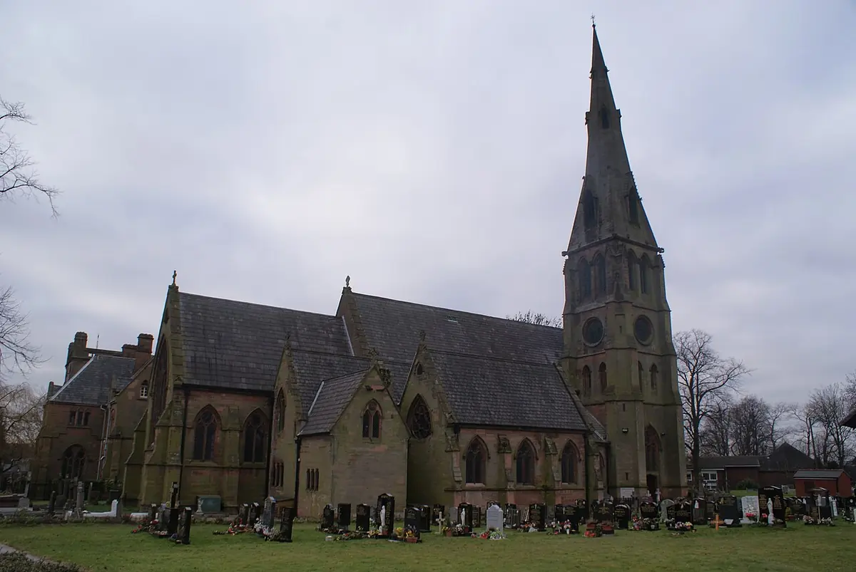 A Victorian Gothic stone church with a tall spire, seen across a churchyard under a gray sky
