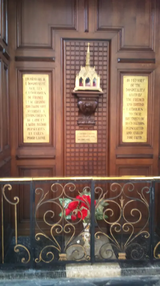 A small reliquary on a carved wooden panel between two brass plaques, above an ornate iron railing, in a church in Douai