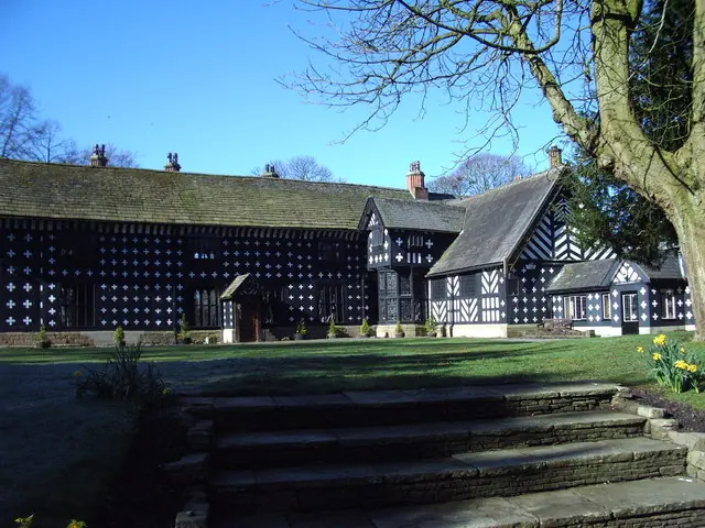 A long black-and-white half-timbered Tudor manor house behind a lawn and stone steps, under a blue sky