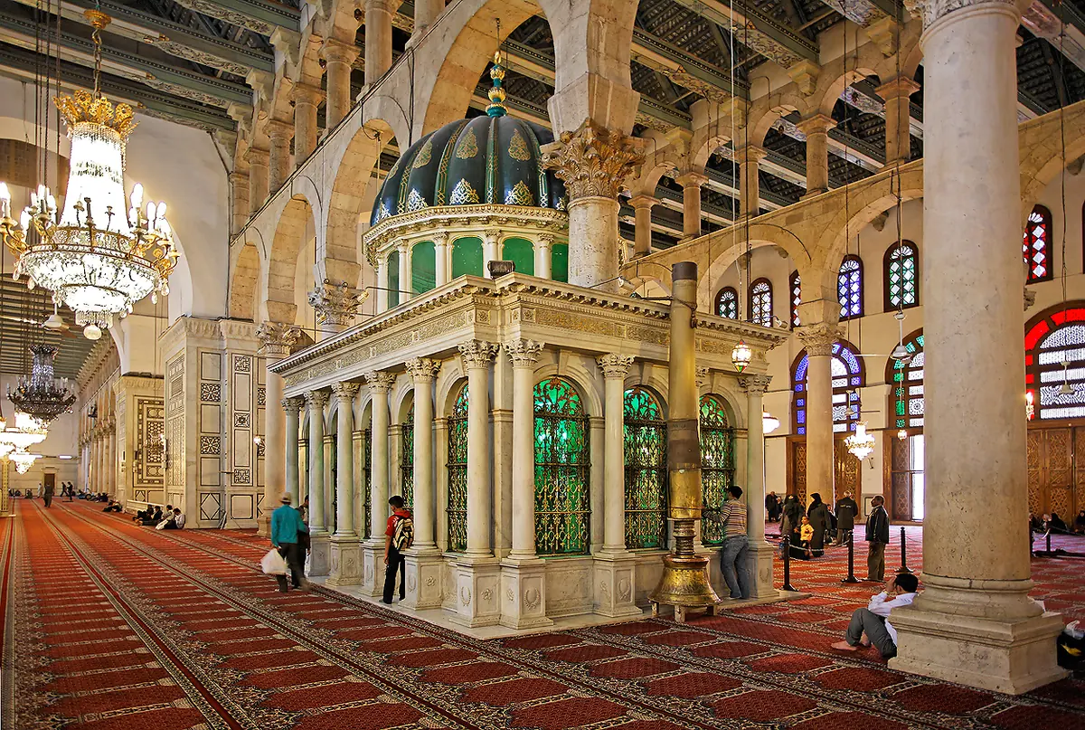 Santuario de mármol con cúpula y ventanas de vidrio verde dentro de la sala de oración alfombrada de la Mezquita de los Omeyas, entre altas columnas