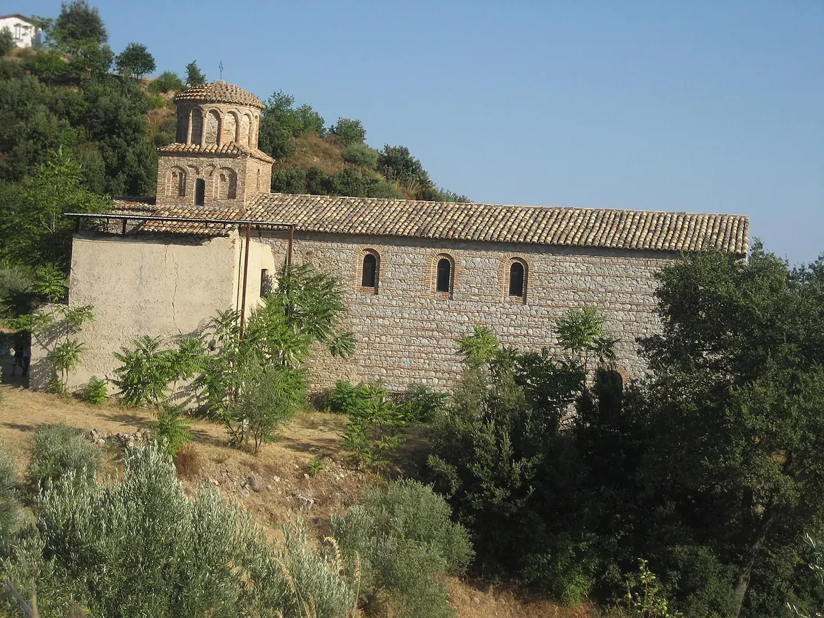 A long stone church with a small round domed tower, rising among olive trees and green shrubs on a hillside in Calabria
