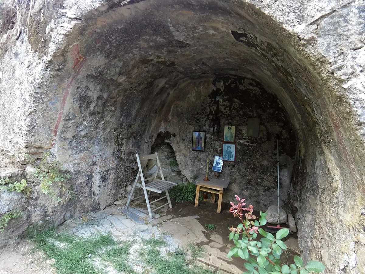 A shallow rock cave in a hillside, with a few small icons, a folding chair and potted flowers inside