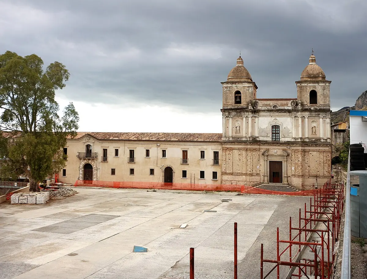 A Baroque church facade with two domed bell towers beside a long former monastery building, on a wide paved square under a cloudy sky