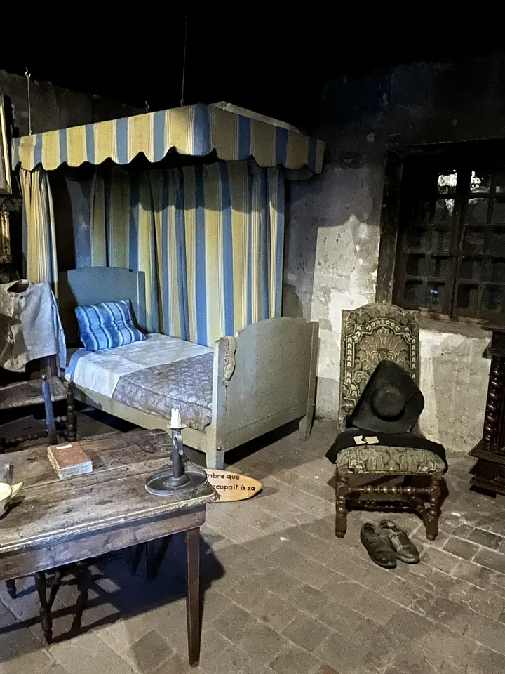 Bare room with a narrow bed under a striped canopy, a worn chair, a small table with a candlestick and a pair of old shoes on the stone floor