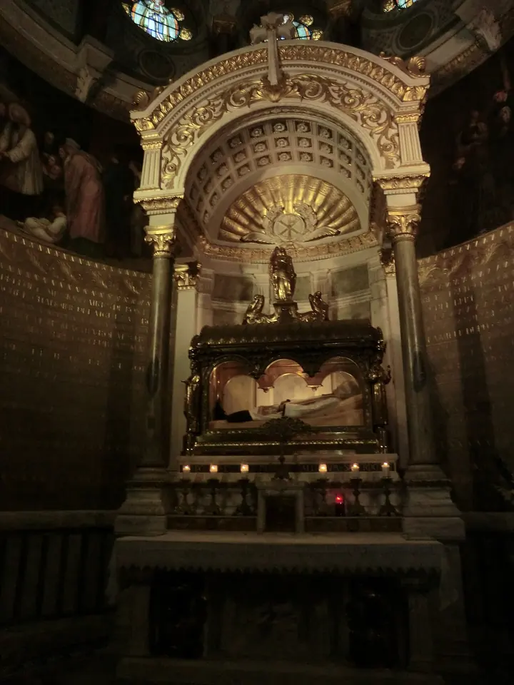 Gilded Baroque-style shrine under a dome, with the body of the Curé of Ars lying in a glass reliquary above an altar lit by candles
