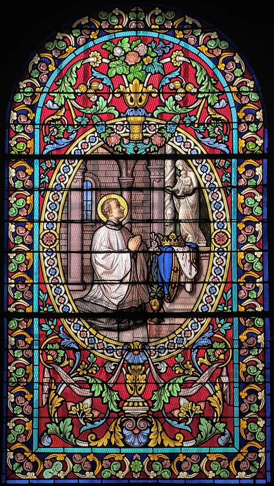 Colorful stained-glass window framed in red and green foliage, showing a priest in a surplice kneeling in prayer before a vision