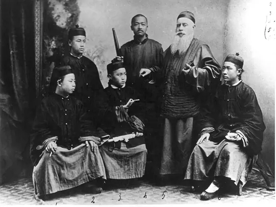 An old studio photograph of a bearded bishop in Chinese dress standing with a Chinese servant and four young Chinese seminarians in long gowns and caps