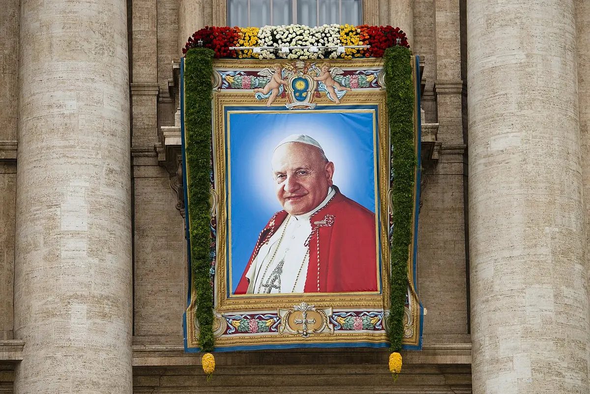 A large woven banner portrait of the smiling pope in a red cape, framed in garlands of flowers and hung between columns of St. Peter's facade
