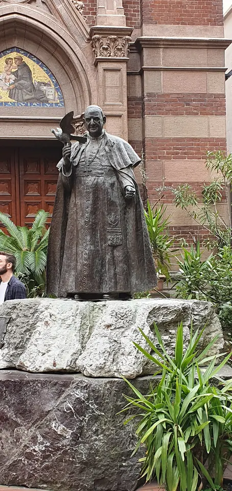 A bronze statue of the pope raising one hand in greeting, standing on a rough stone base in the courtyard of a red brick neo-Gothic church
