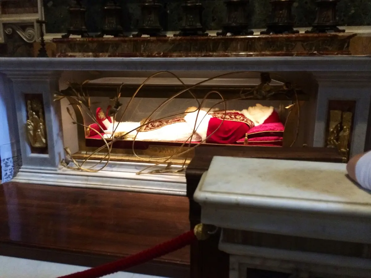 A glass and gilded bronze case beneath a marble altar, holding the body of the pope dressed in red and white vestments