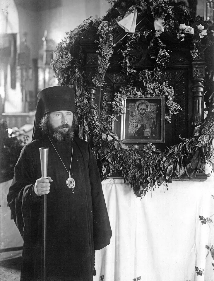 A black-and-white photograph of a bearded bishop in a black klobuk holding a tall staff, standing beside an icon of St. Nicholas framed in greenery