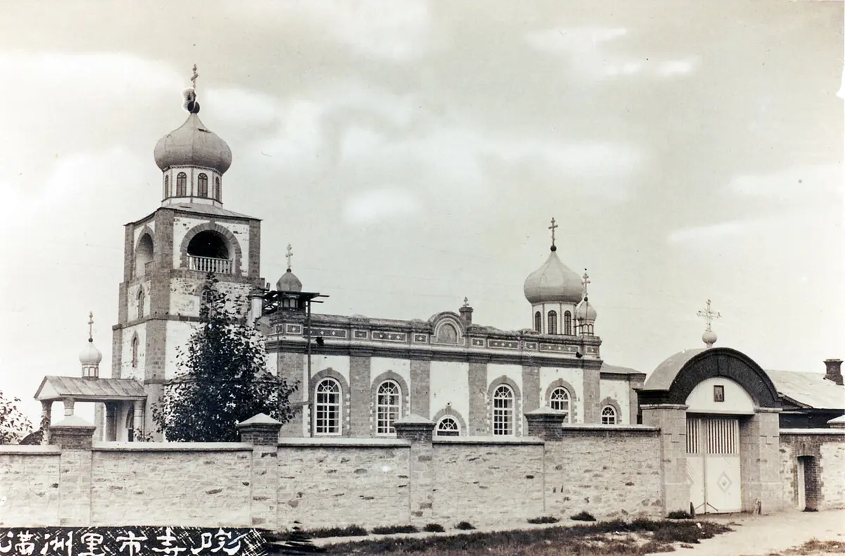An old photograph of a large church with a bell tower and onion domes behind a long wall in a flat, treeless town