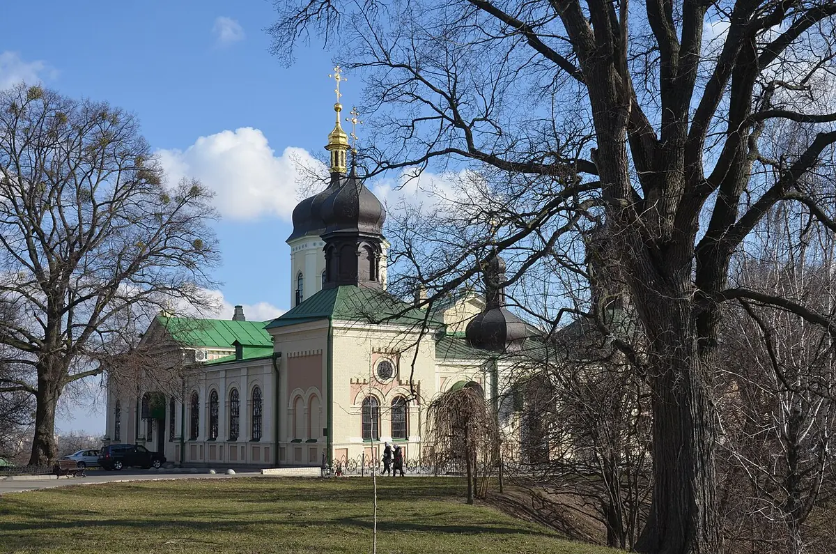 A pale yellow church with a dark onion dome and green roofs among bare trees in early spring