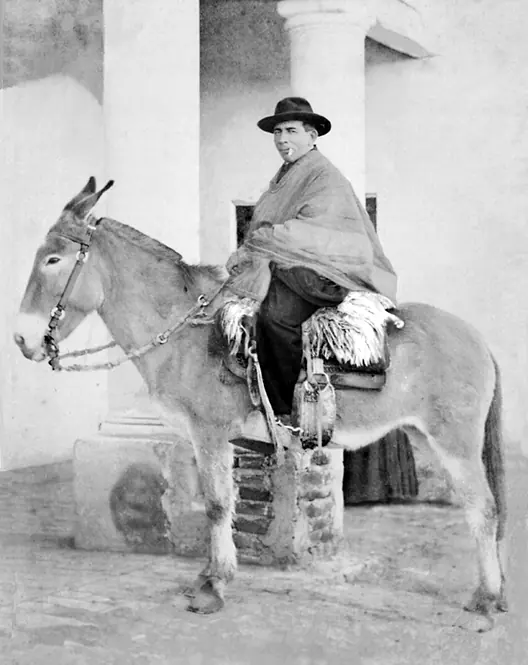 An old photograph of a heavyset priest in a hat and poncho sitting on a mule in front of a building with columns