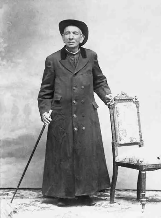 An old full-length photograph of an elderly priest in a long dark coat and a wide-brimmed hat, leaning on a cane beside a chair