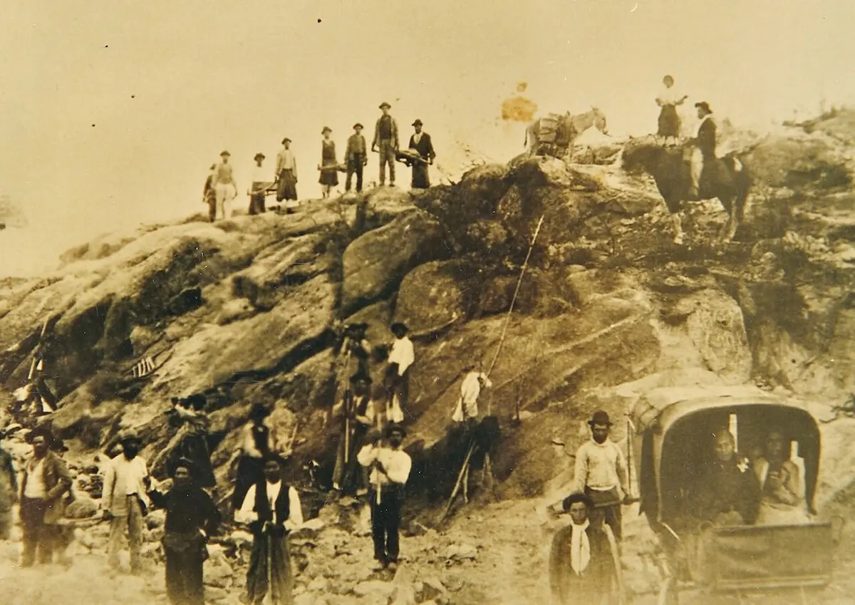 An old sepia photograph of a crowd of men with tools and a cart working on a road cut into a rocky hillside
