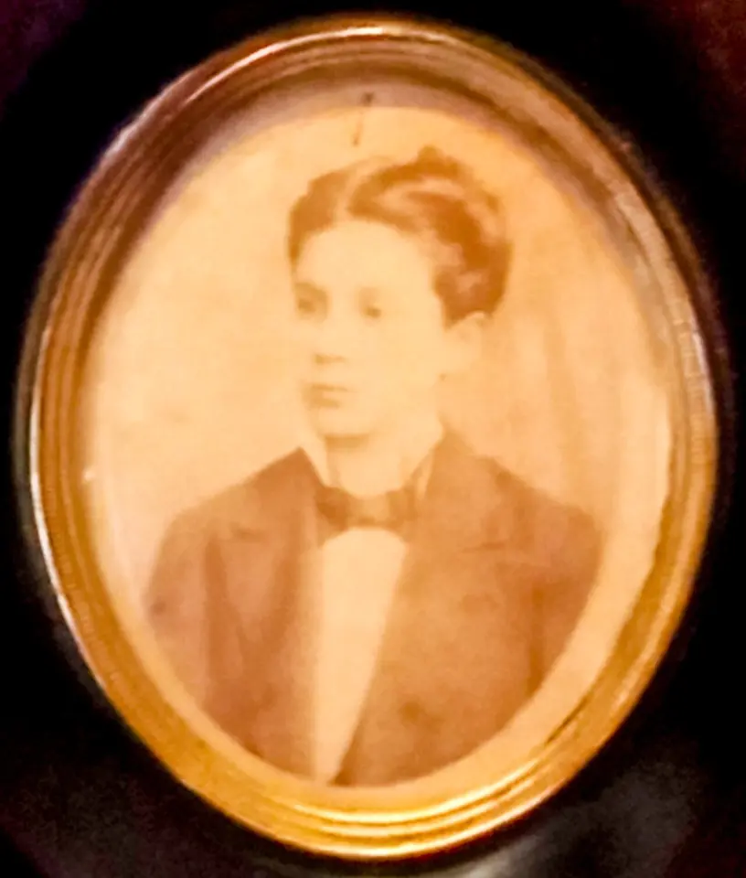 Faded oval sepia photograph of a teenage José Gregorio Hernández with wavy hair, in a dark jacket and bow tie, in a gilt frame