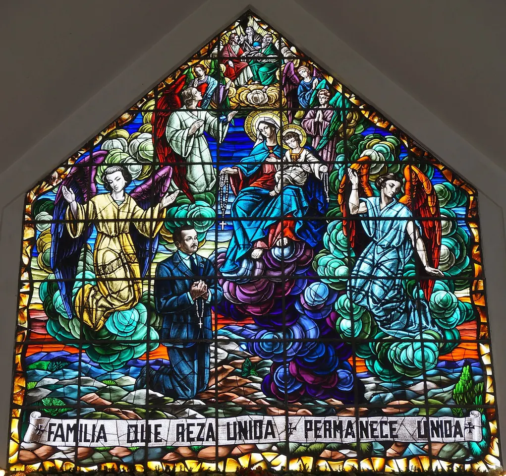 Bright stained-glass window showing Dr. Hernández in a dark suit kneeling with a rosary beneath the Holy Family and angels, with the words "Familia que reza unida permanece unida"