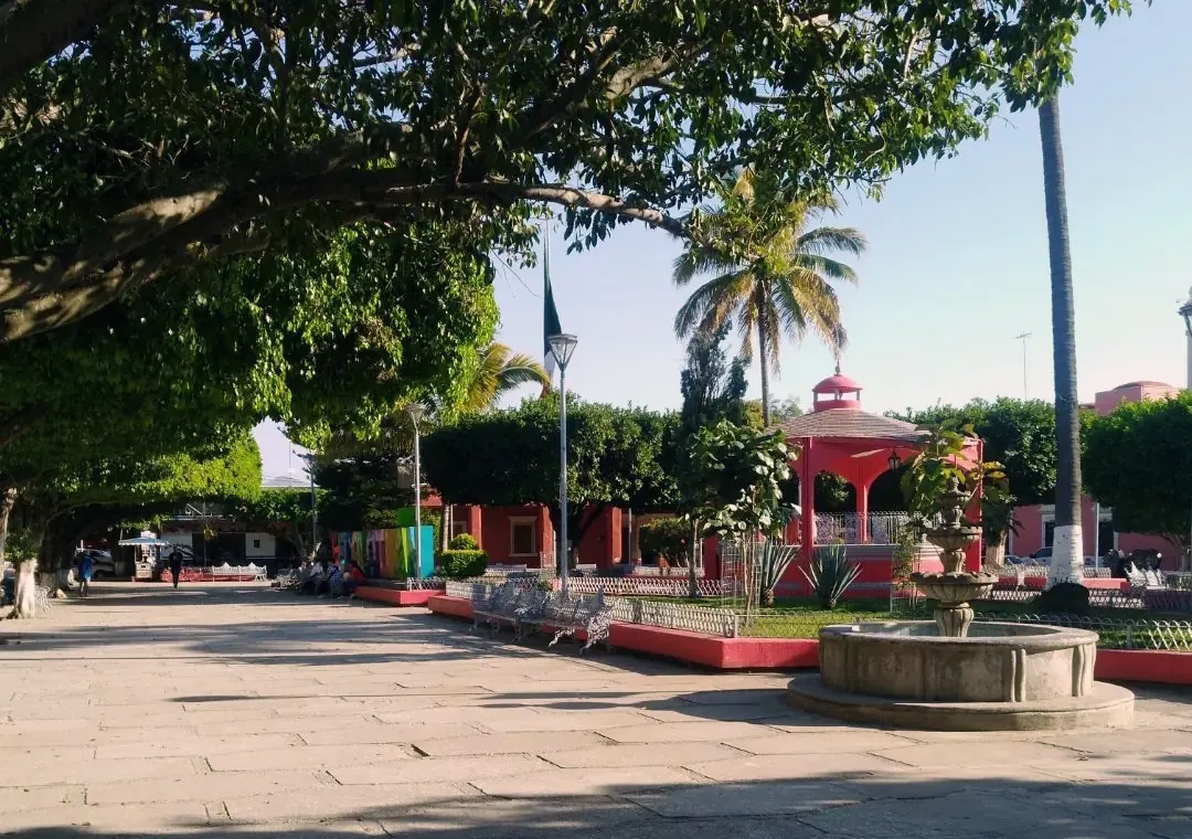 A shady town square in Mexico with a stone fountain, white iron benches, a red bandstand and palm trees