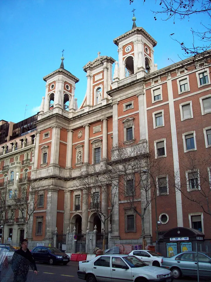 The red brick and stone facade of a church with two bell towers on a city street in Madrid, with parked cars in front