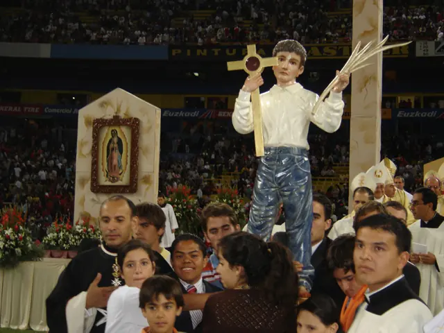 A statue of a boy in a white shirt and blue trousers holding up a cross and a palm branch, carried through a crowded stadium beside an image of Our Lady of Guadalupe
