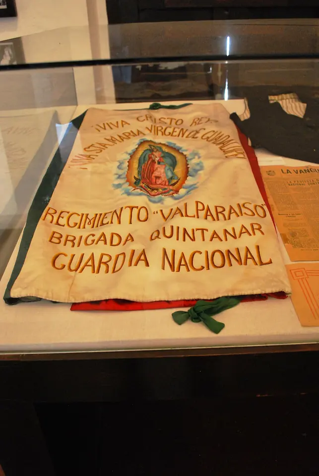 An old cream-colored banner in a display case, painted with an image of Our Lady of Guadalupe and the words "Viva Cristo Rey" and "Regimiento Valparaíso, Brigada Quintanar, Guardia Nacional"
