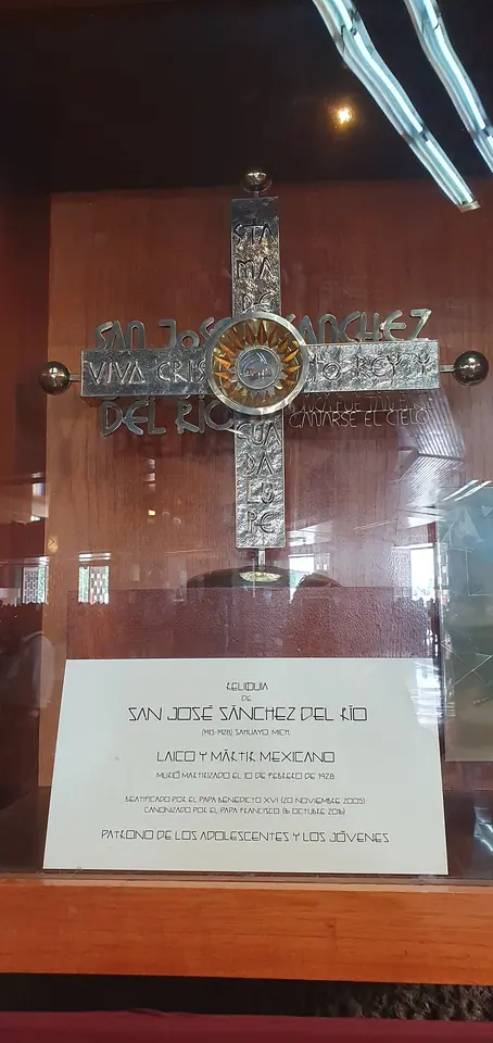 A silver cross reliquary with a round relic case at its center, inscribed "Viva Cristo Rey," in a glass case above a label naming St. José Sánchez del Río