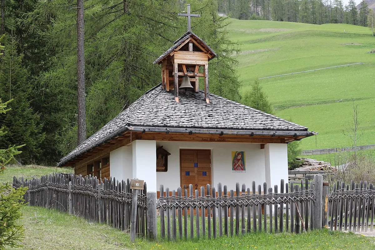 A small white chapel with a shingled roof and a wooden bell turret, behind a wooden fence, in a green Alpine meadow with larch trees
