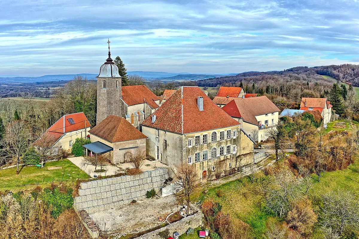 A small French hill village with a stone church and bell tower among red-roofed houses, wooded hills and farmland behind