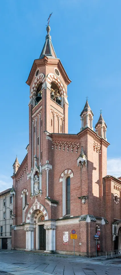 Tall red-brick church with white stone bands, a pointed bell tower and a statue above the doorway, on a street corner in Asti